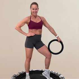 Woman in athletic wear standing on a trampoline holding an exercise ring against a beige background